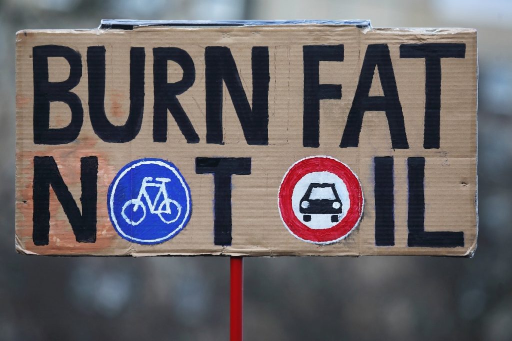 Color photo of a protest sign. “BURN FAT NOT OIL” is painted in big black letters on a rectangular cardboard. The “O” in “NOT” is replaced by a painting of a blue traffic sign featuring a small white bicycle icon in its middle. The O” in “OIL” is replaced by a painting of a red and white traffic sign featuring a black car icon in its center.