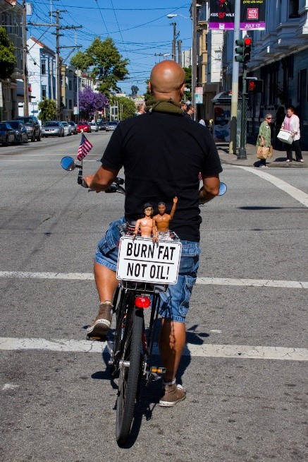 Color photo of a cyclist. The picture is taken from behind and shows the back of a cyclist, wearing a black t-shirt and blue jeans, standing on a street in front of a red traffic light. The bicycle has rear mirrors on both sides; a small American flag is attached to the left one. On the back of the bike in the picture’s center is a numberless license plate that reads “BURN FAT – NOT OIL!” Two male, shirtless Ken dolls, one white and one Black, are attached to the upper edge of the plate.